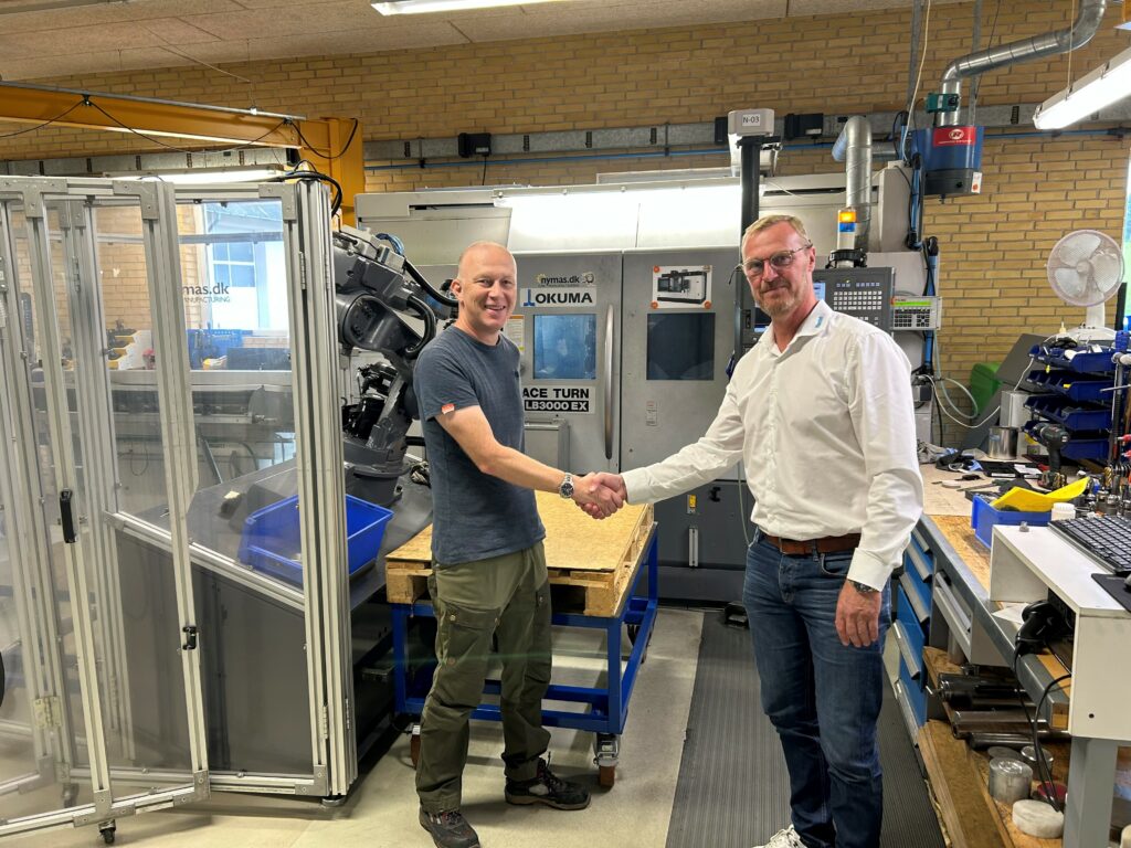 Anders Larsen fra Flextek og fabrikschef Torben Pryds fra BJ Gear giver hinanden hånden foran en Okuma-maskine hos BJ-Gear A/S, omgivet af automatiseringsudstyr og robotcelle.