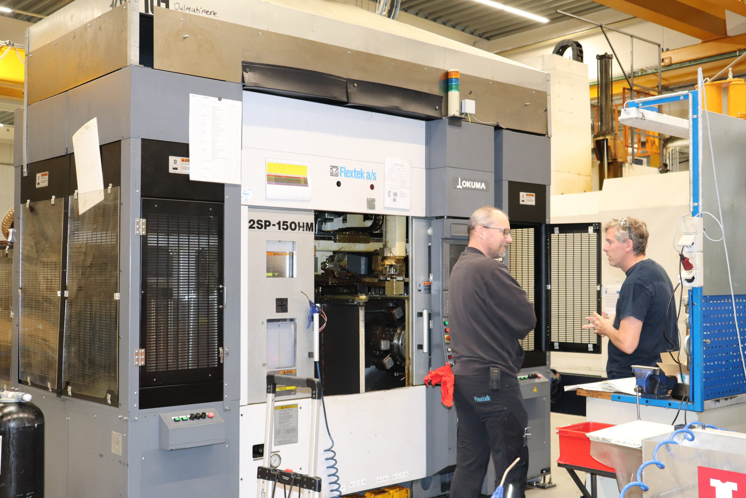 To teknikere i dialog foran Okuma 2SP-150HM CNC-maskine i produktionsmiljø hos Niebuhr Gears.